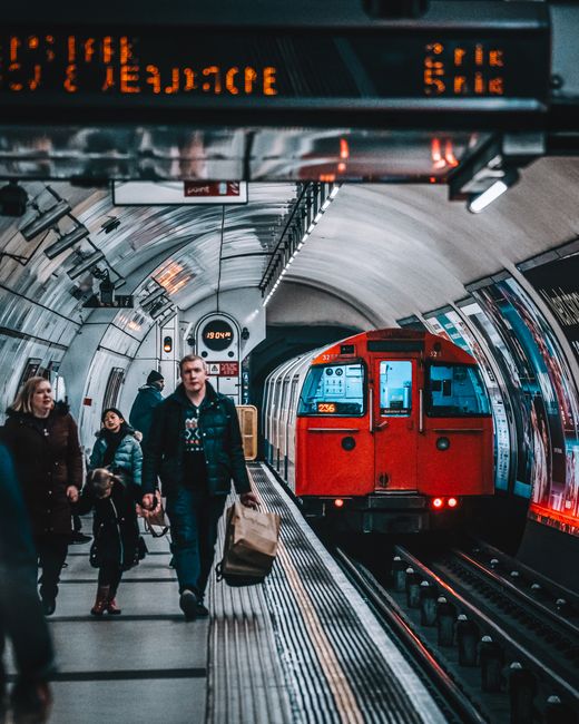 A busy London underground station with a red train approaching the platform. Several passengers, including a man holding a paper bag and a woman with a young girl, are walking along the tiled platform