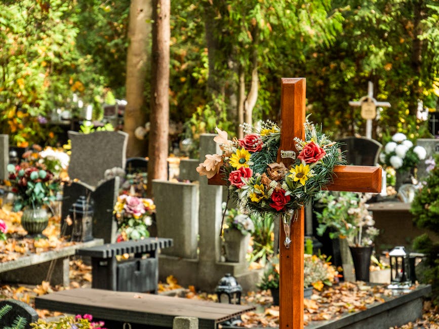 A wooden grave cross adorned with a floral arrangement consisting of red roses, yellow daisies, and pink lisianthus, surrounded by green foliage. The cross is set in a cemetery with other headstones a