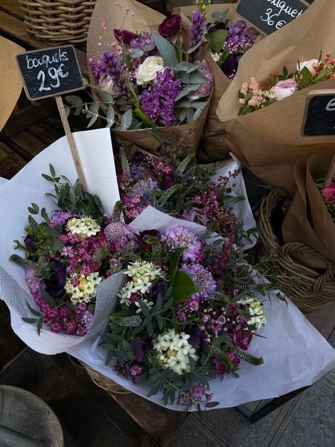 A collection of fresh floral bouquets arranged with various purple, white, and pink flowers, including lavender, alstroemeria, and waxflowers, wrapped in brown kraft paper and white tissue paper. The 
