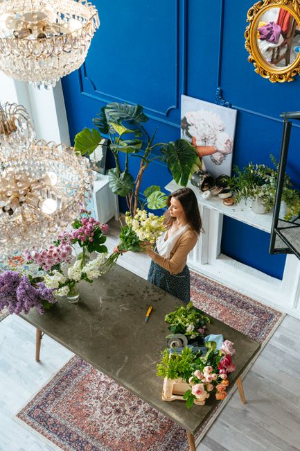 A woman with long brown hair, dressed in a beige top and dark patterned skirt, is arranging a bouquet of fresh flowers on a grey table inside a brightly lit florist shop. The bouquet features white an
