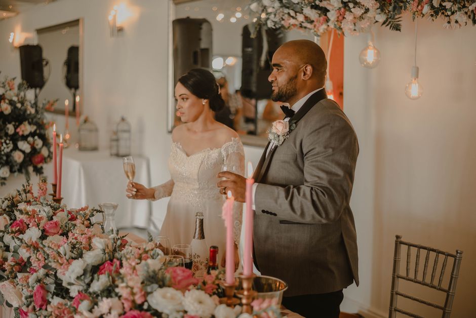 A man and woman at a wedding reception stand beside a table adorned with a large, elaborate floral arrangement featuring pink, white, and peach roses, complemented by greenery and delicate filler flow