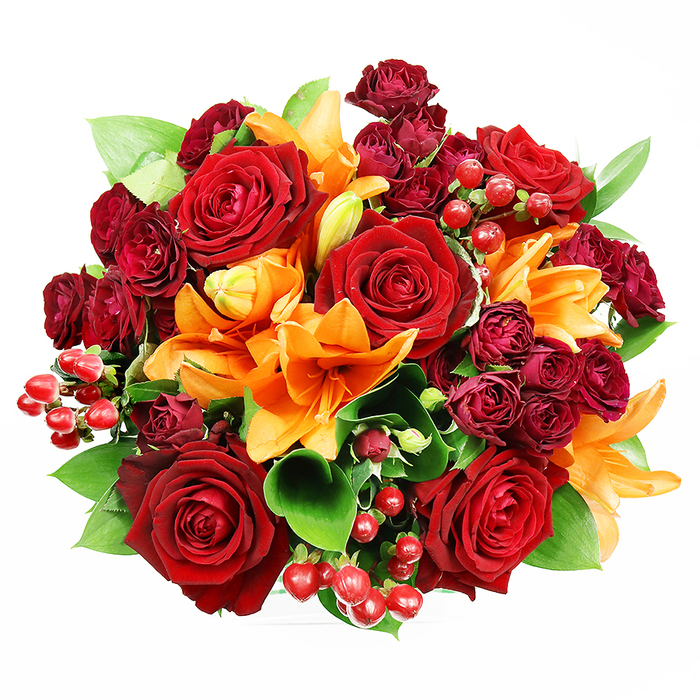 Captured overhead on a pristine white surface, this luxurious circular bouquet is a study in warm, opulent contrast - deep red roses paired with bright orange Asiatic lilies and rich, wine-colored spray roses. The largest roses sit prominently at the centre, their velvety petals unfurling in layered spirals that suggest long-stemmed romance and a comforting floral perfume. Bright orange lilies punctuate the circle with trumpet-shaped elegance; some reveal dark speckling and prominent stamens while others remain in graceful bud form, lending a sense of unfolding life. Intermittent clusters of small burgundy spray roses add concentrated colour and texture, while clusters of glossy red hypericum berries sparkle like tiny rubies, providing lively, reflective highlights. Lush green ruscus or salal leaves weave between blossoms, their broad glossy surfaces giving a cooling, structural frame to the warm palette. The lighting is soft and even, modeled to show the depth of each petal and the sheen on the berries without harsh shadow, evoking the quiet morning light you might notice near Cockfosters Tube or while walking the paths of Trent Park. As a local craftsman's piece from Florist Cockfosters, this bouquet is designed to convey devotion and celebration - ideal for anniversaries, romantic gestures, or as an elegant addition to autumnal home décor - and it reads as both richly expressive and carefully balanced.